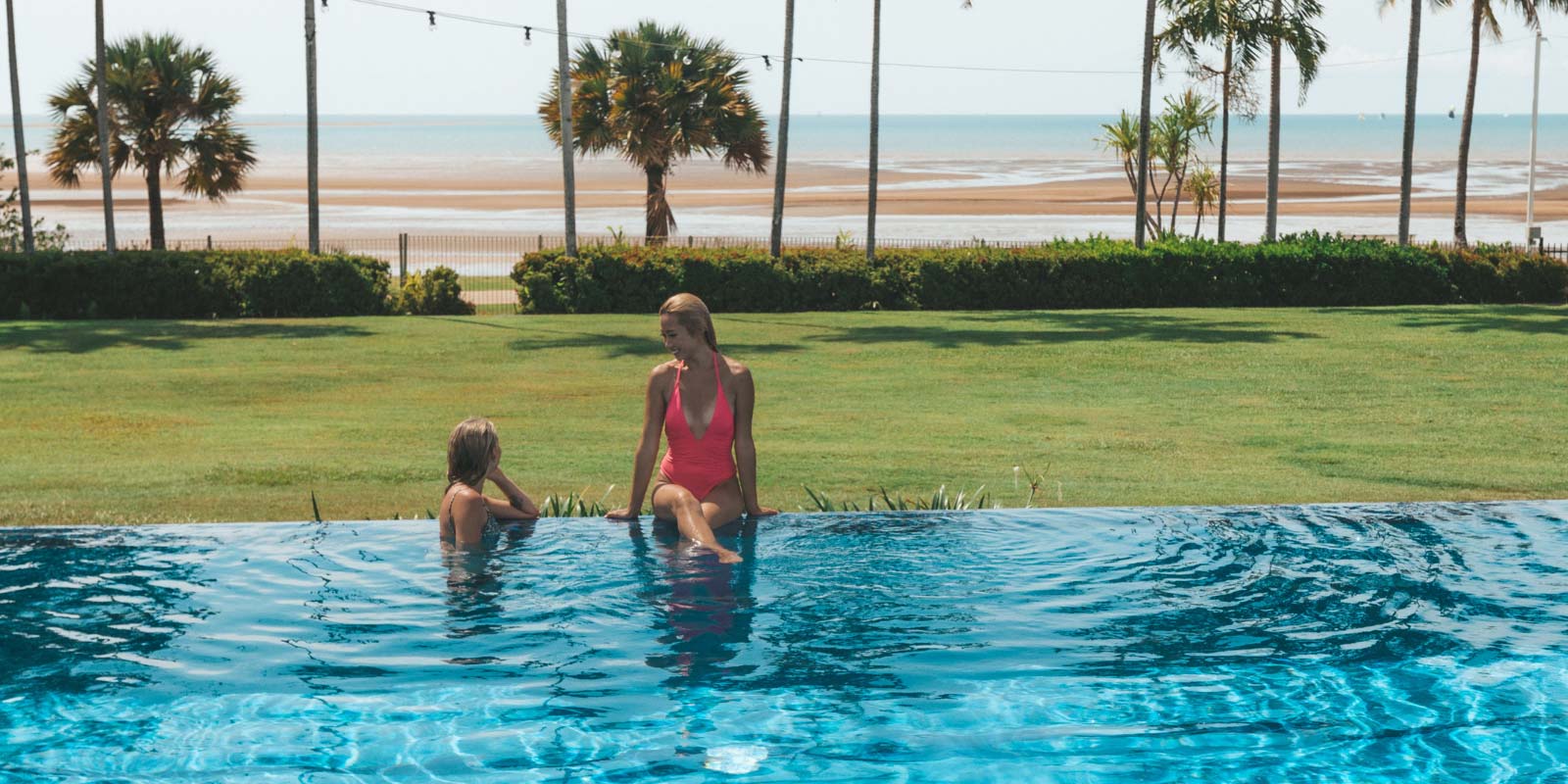 Two women sitting at the edge of the infinity pool looking out at the lawn and beach shore.